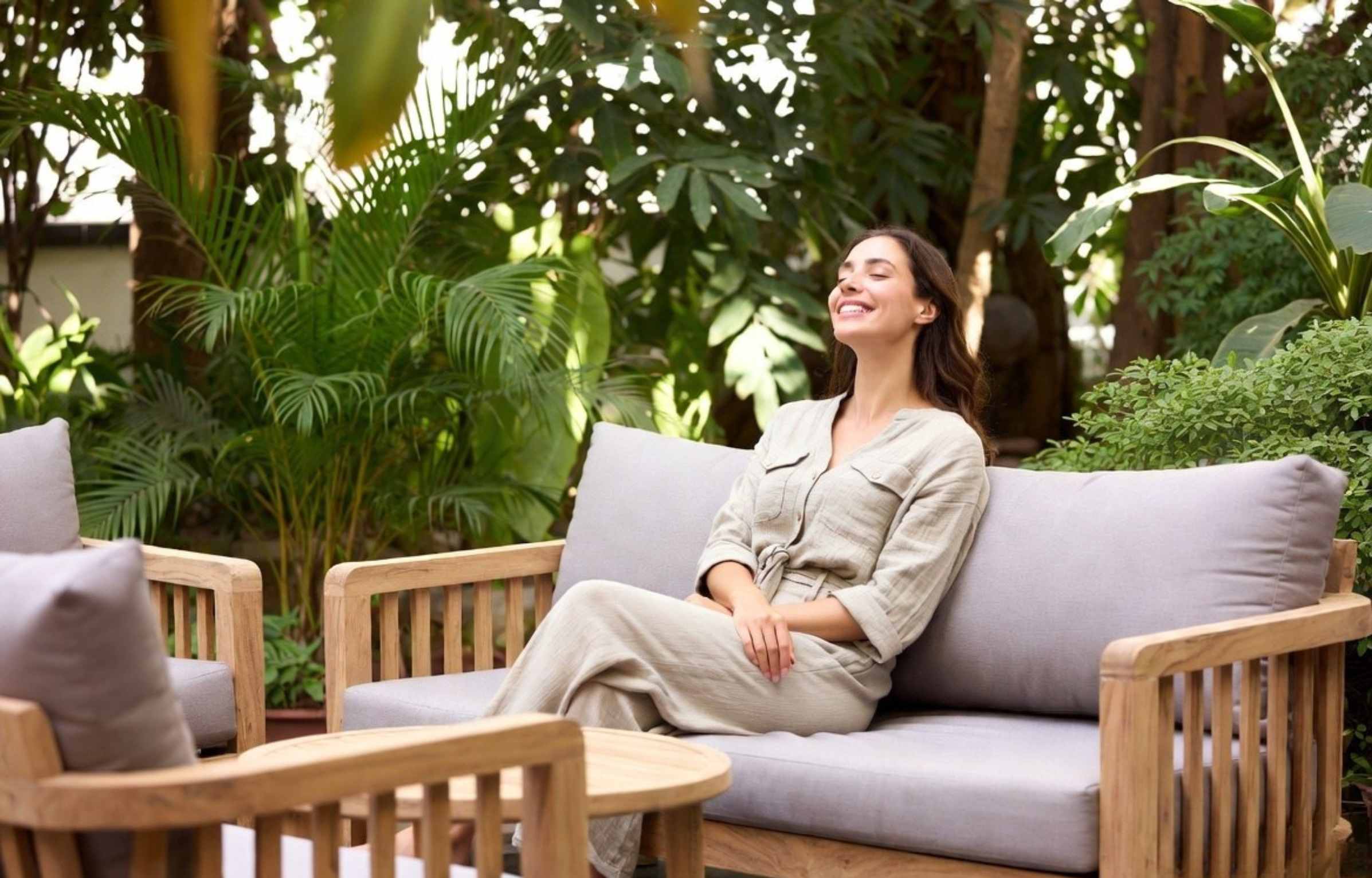 Woman relaxing on a teak outdoor sofa set surrounded by lush garden greenery.