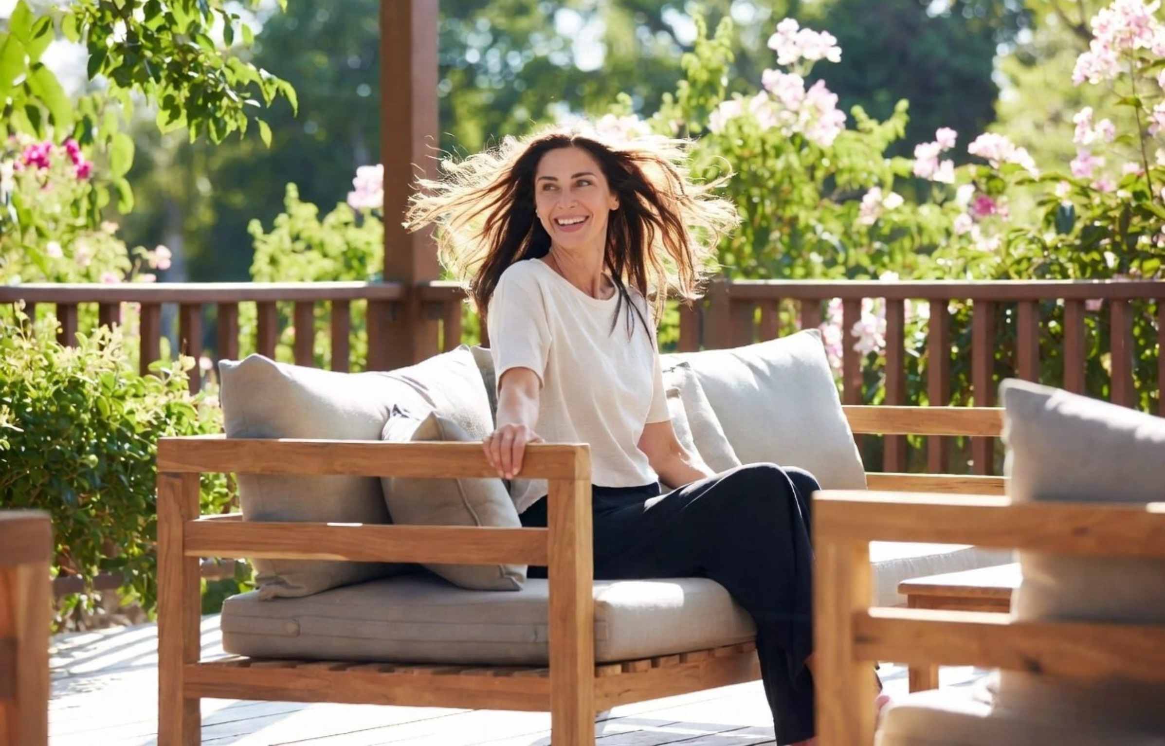 Woman sitting on outdoor sofa sets with light cushions and flowers in a teak garden setting.