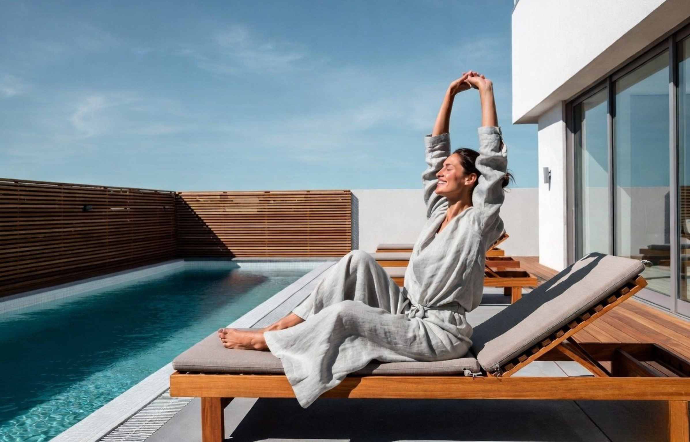 Woman stretching on a teak sun lounger near a modern pool.
